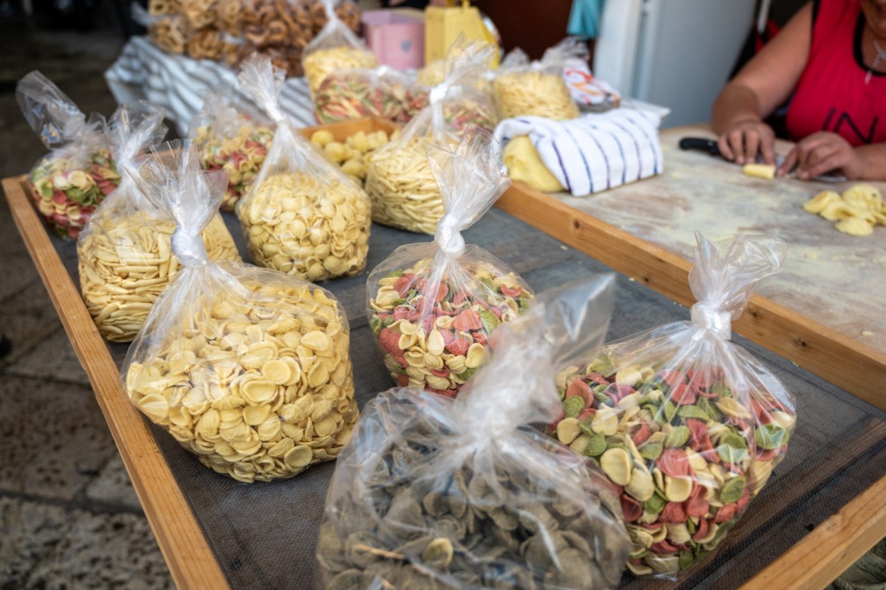 Several transparent bags filled with pasta lie on a dark sieve. In the background, a person sits at a table making more pasta.