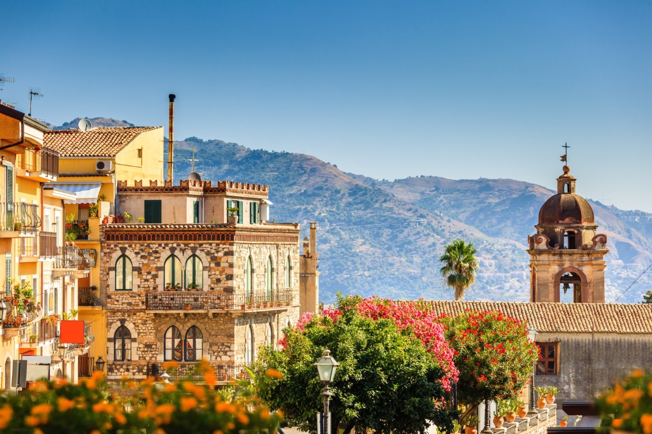 Buildings, church tower, flowering plants and trees, mountains in the background 