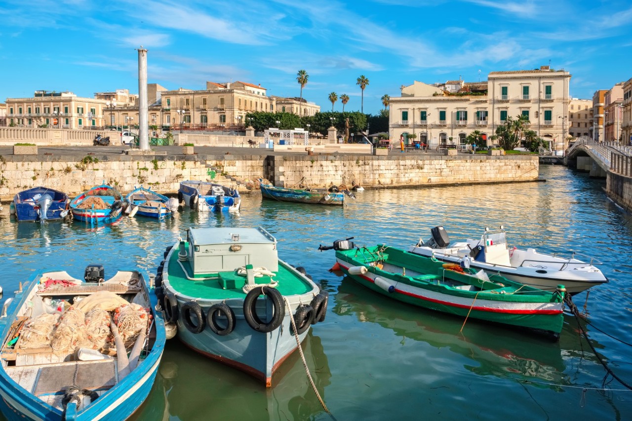 Fishing boats docked on calm water, quay wall, buildings, a column, trees and palm trees in the background 