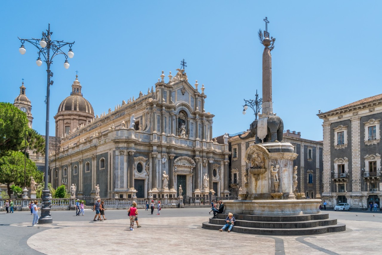 Large square with a fountain at the centre and pedestrians, flanked by a two-storey baroque cathedral, behind it two domes and next to it magnificent town houses 