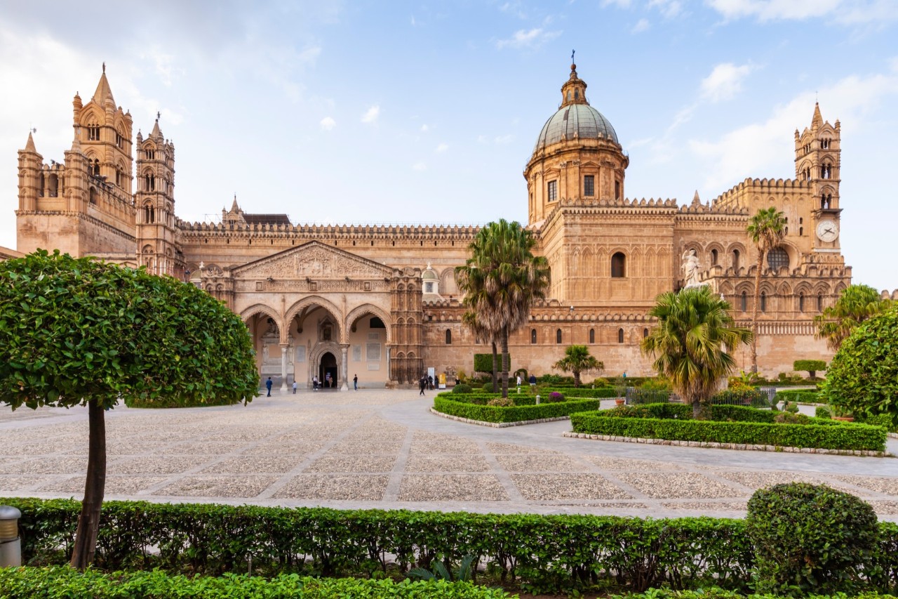 Cathedral made of light-coloured sandstone with an almost deserted large front courtyard, palm trees, low hedges  