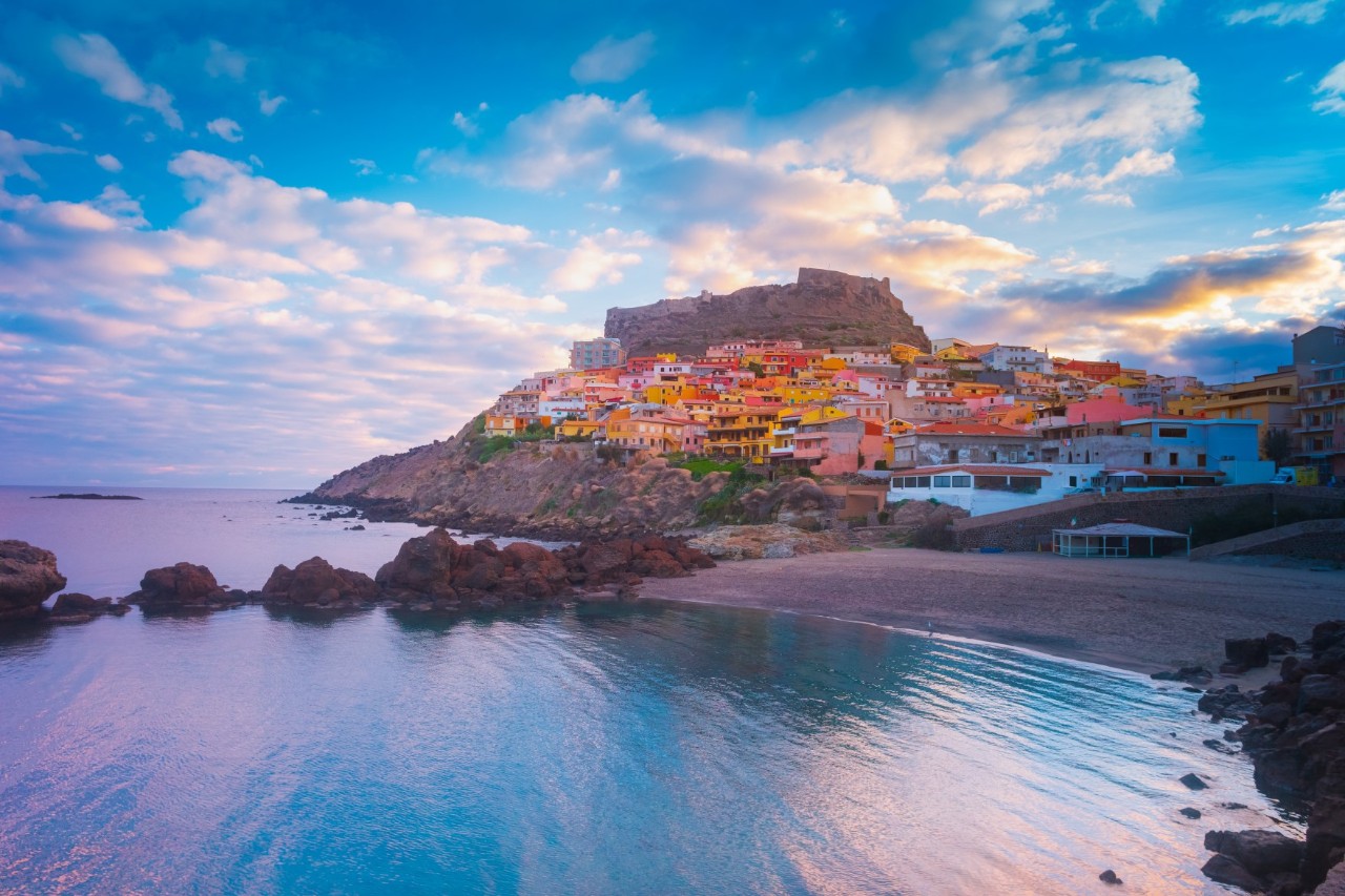picturesque coastal town. Colourful houses nestle against a rocky hill on which a medieval castle is enthroned. In the foreground is a quiet beach and the blue-purple sea in the evening light.