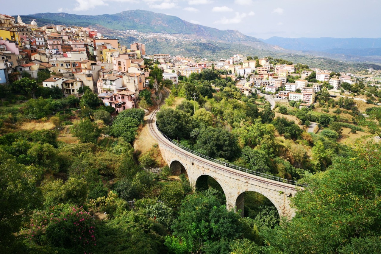 The picture shows a hilly landscape with a village on a slope consisting of colourful buildings. In the foreground, a stone viaduct runs through lush green vegetation. Mountains rise up in the slightly misty background.