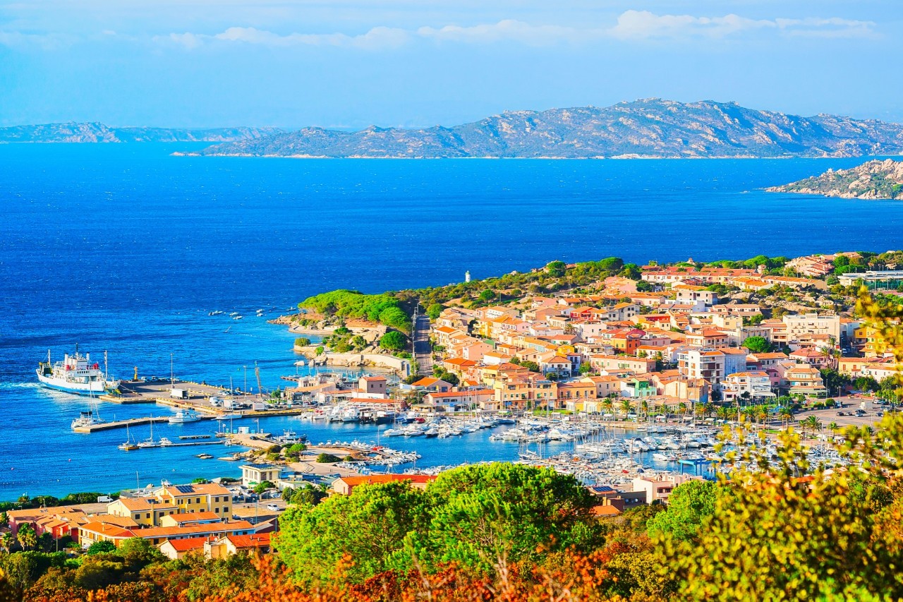 View from above of a coastal town with a harbour, surrounded by turquoise blue sea and wooded hills. In the background, islands or headlands stretch across the water. 