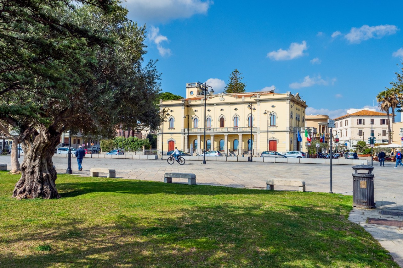 The picture shows a large, quiet square. In the foreground is a well-tended green area with an old tree. In the background is a classic, light-yellow building. 