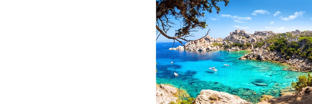 Picturesque bay with turquoise-blue water in which a few small boats are moored. The bay is surrounded by rocky cliffs and dense green vegetation. 