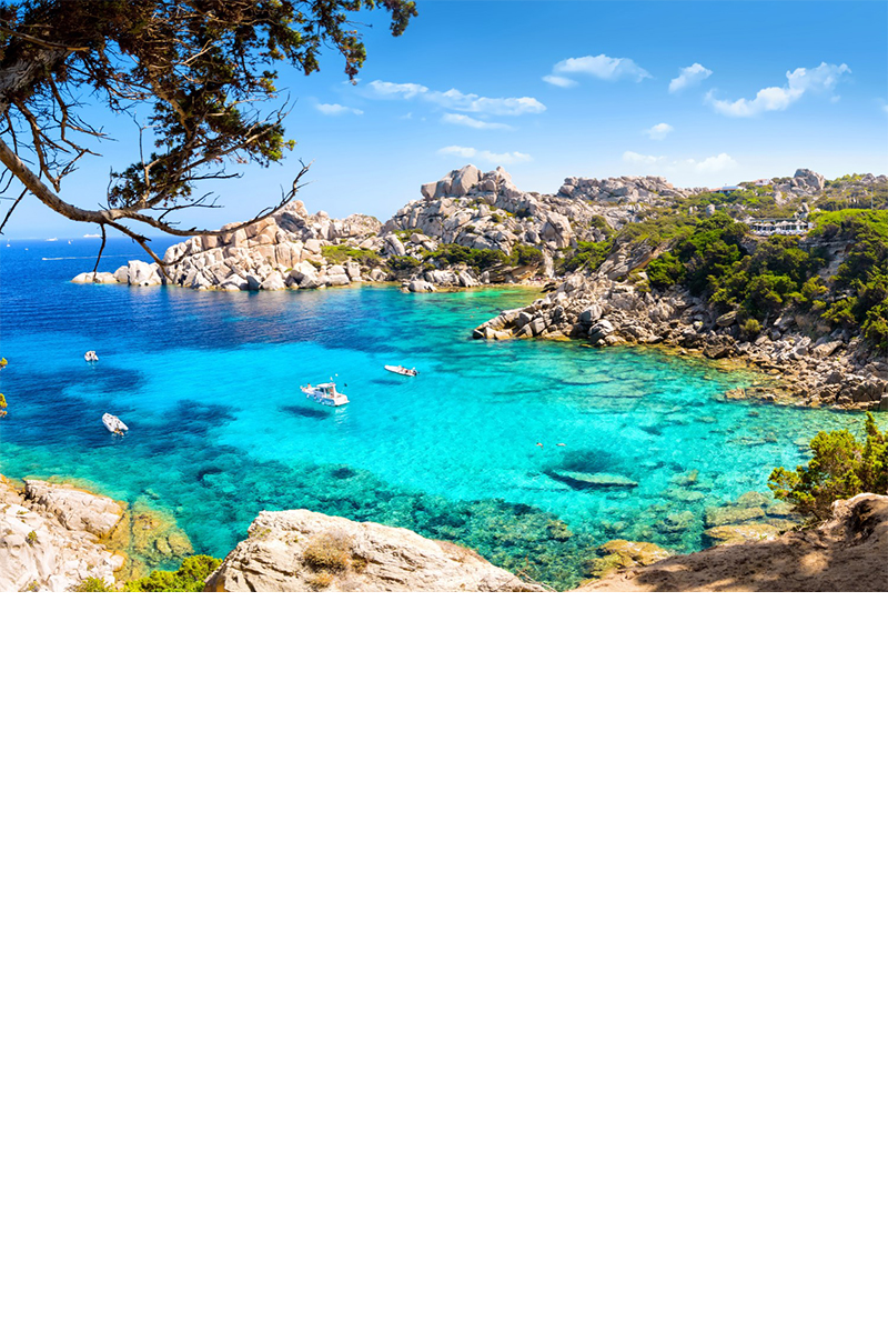 Picturesque bay with turquoise-blue water in which a few small boats are moored. The bay is surrounded by rocky cliffs and dense green vegetation. 