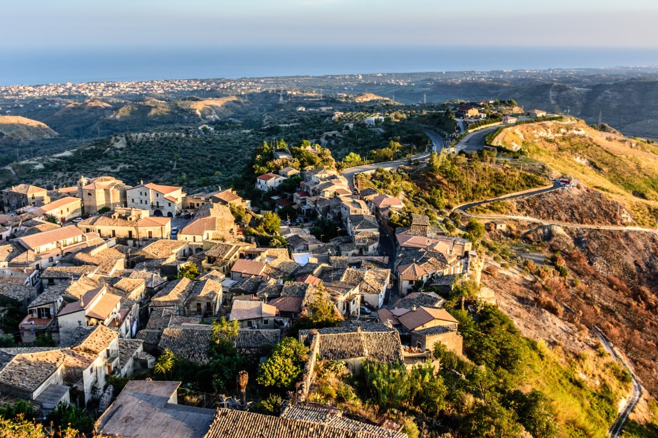 View from above of a village with houses built close together and tiled roofs on a hill. In the background, a wide landscape extends to the sea on the horizon.