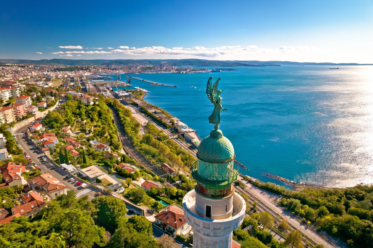 Bird’s eye view of the city with buildings and trees directly by the sea and an angel-like statue on the tower in the foreground.