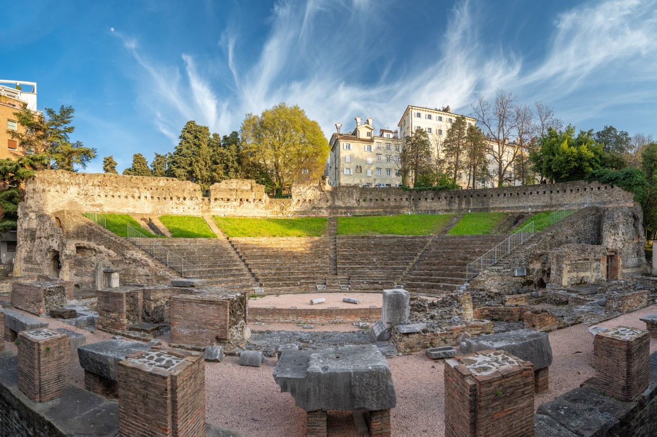 View of the remains of a semi-circular Roman amphitheatre with trees and a white building in the background. 