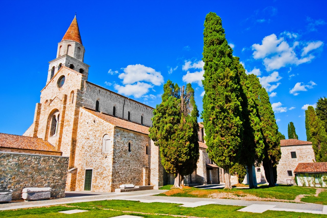 Large church building with several outbuildings, deserted square and trees on a sunny day.