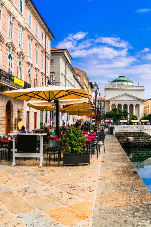 View of Trieste city centre: elegant buildings and palaces along a canal in which they are reflected, street cafés with parasols and plants. 