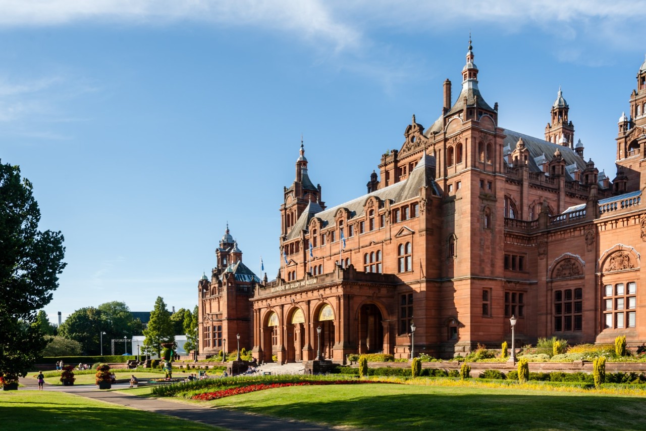 On the right-hand side of the picture is an impressive Victorian-style building. The facade is made of red sandstone with ornate towers and elaborate details. Well-kept gardens with flowerbeds can be seen in front of the building.