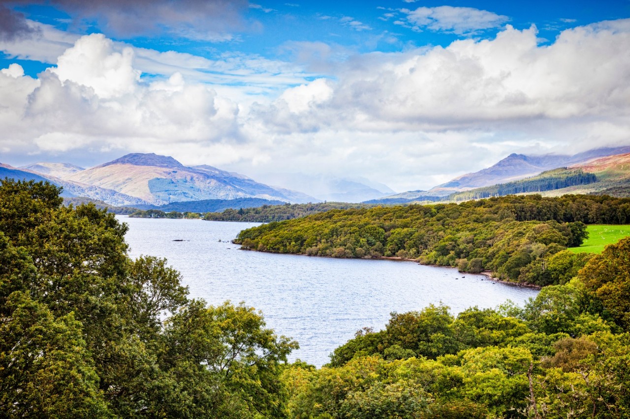 The picture shows an eye-catching landscape with a large lake surrounded by densely forested shores and rolling hills. More distant mountains rise up in the background. The sky is overcast and blue.