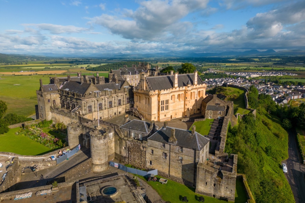 View from above of a majestic, historic castle on a hill, surrounded by green countryside. The castle consists of old stone walls, towers and a striking yellow building. There is a small town in the distant background on the right.