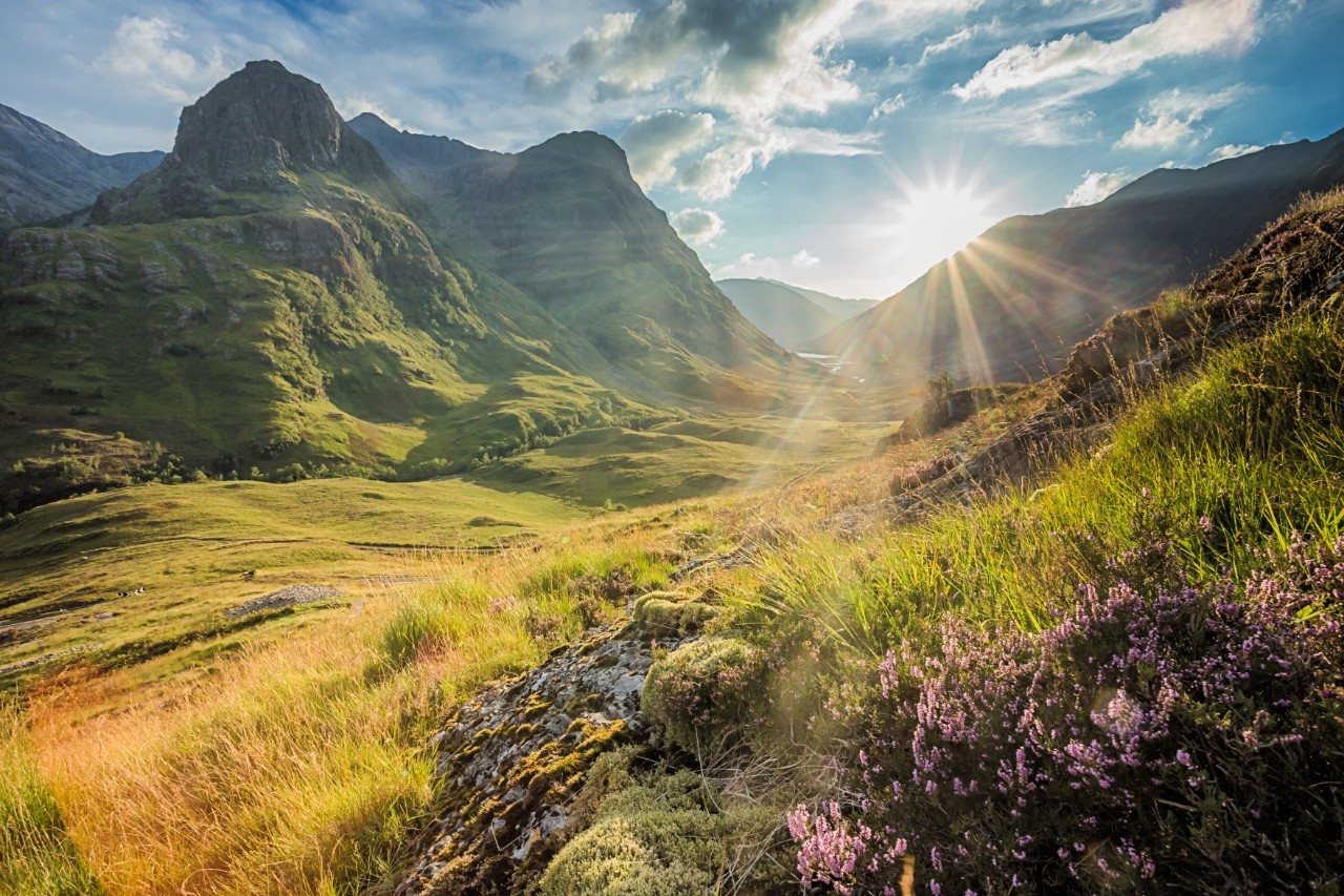 The picture shows an idyllic mountain landscape with green hills and rocks. The sun emerges from behind a rock and shines into the valley in the foreground. Grasses and heather can be seen in the foreground, while steep mountains and a valley characterise the background scene.