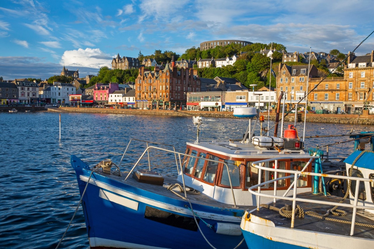 View from the water of a harbour town with buildings of different sizes and light, sand-coloured and colourful facades. Boats are in the foreground. In the background, a hill with historic buildings and a round ruin on the hill can be seen.