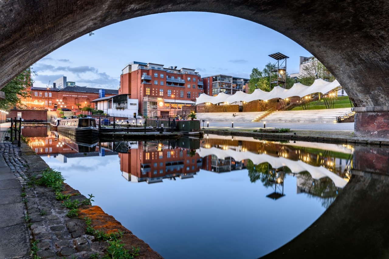 Bridgewater Canal photographed through a bridge arch, reflection of the arch in the water, view of small harbor, seating along the canal and buildings in the background 