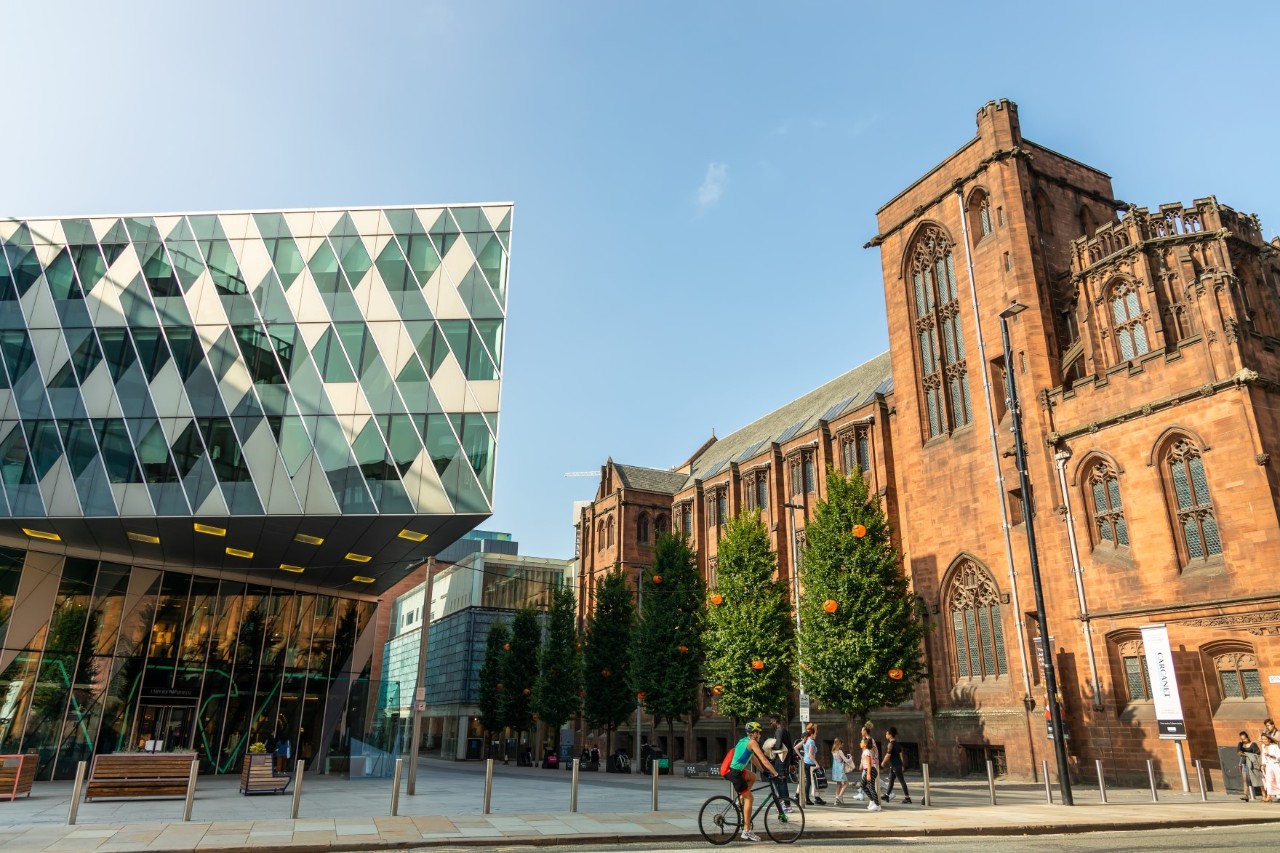 Modern building with large glass façade next to a Gothic building, in the foreground pedestrians, a cyclist and seating 