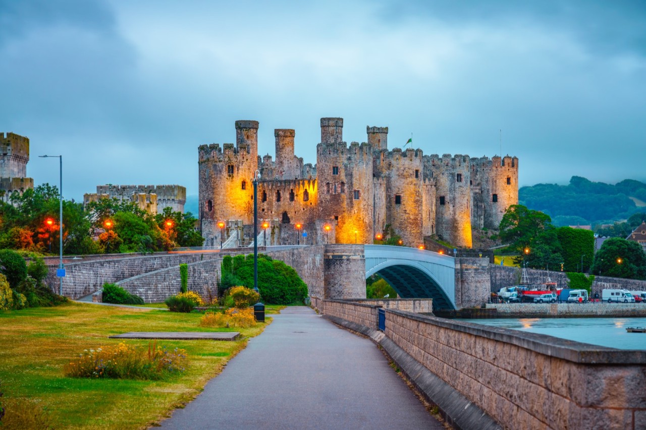 : Illuminated castle at dusk, bridge, stone wall, pedestrian path and river in the foreground © Pawel Pajor/stock.adobe.com
