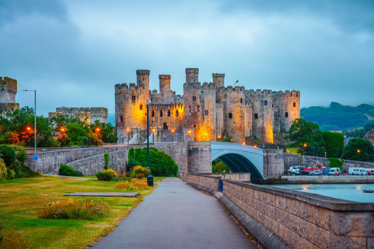 : Illuminated castle at dusk, bridge, stone wall, pedestrian path and river in the foreground 