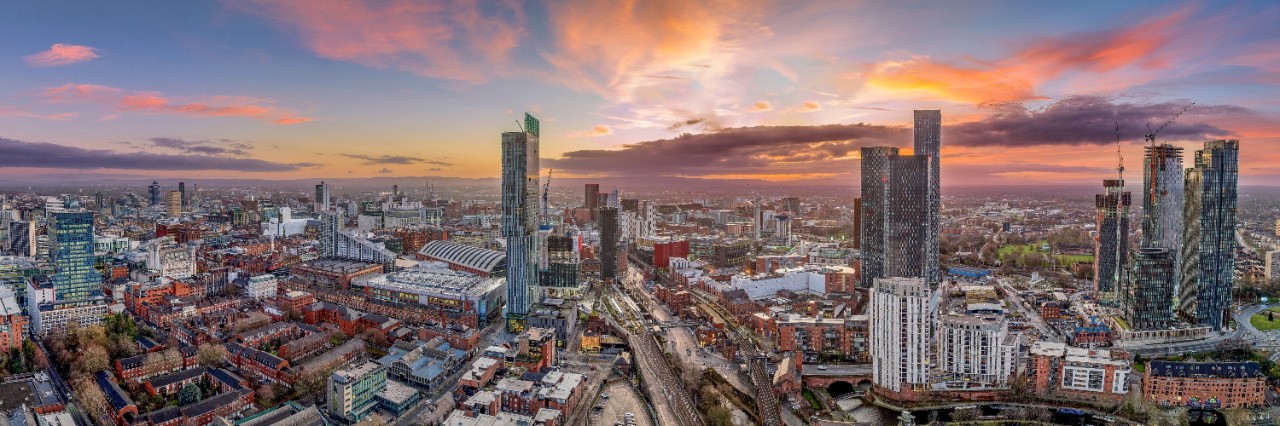 Wide view over the rooftops of Manchester, mix of historic and modern buildings, occasional skyscrapers, sunset. © Chris/stock.adobe.com