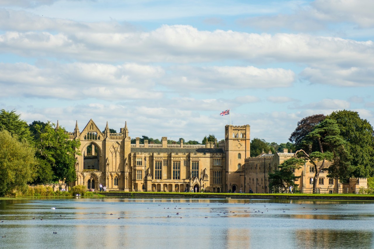 The image shows a beige-coloured historic building in Gothic style with large windows and a British flag on a tower. The complex is surrounded by trees and green countryside. A large lake stretches out in the foreground. 