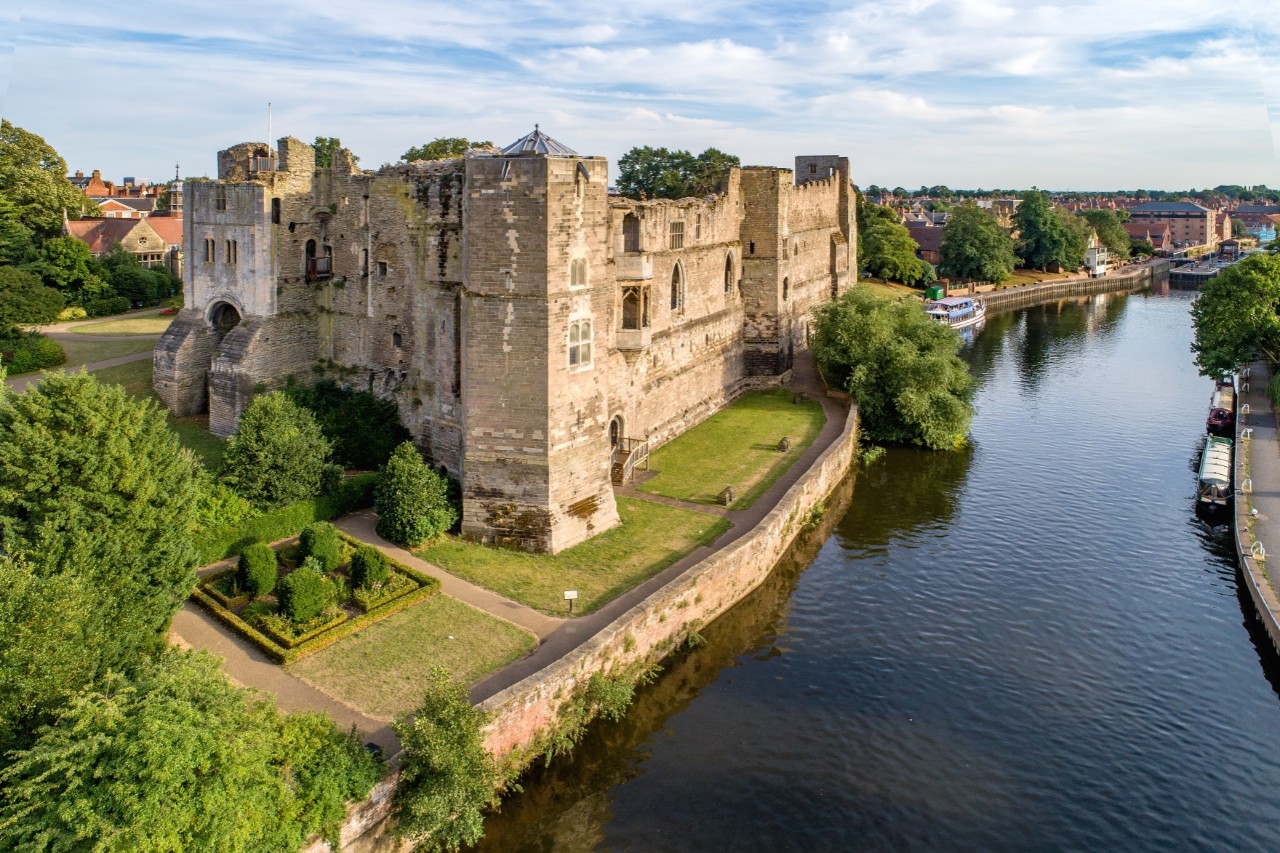 The image shows historic stone castle ruins located on the banks of a peaceful river. The castle walls are surrounded by green spaces and well-tended gardens. Houses can be seen in the background and there are several boats in the river.