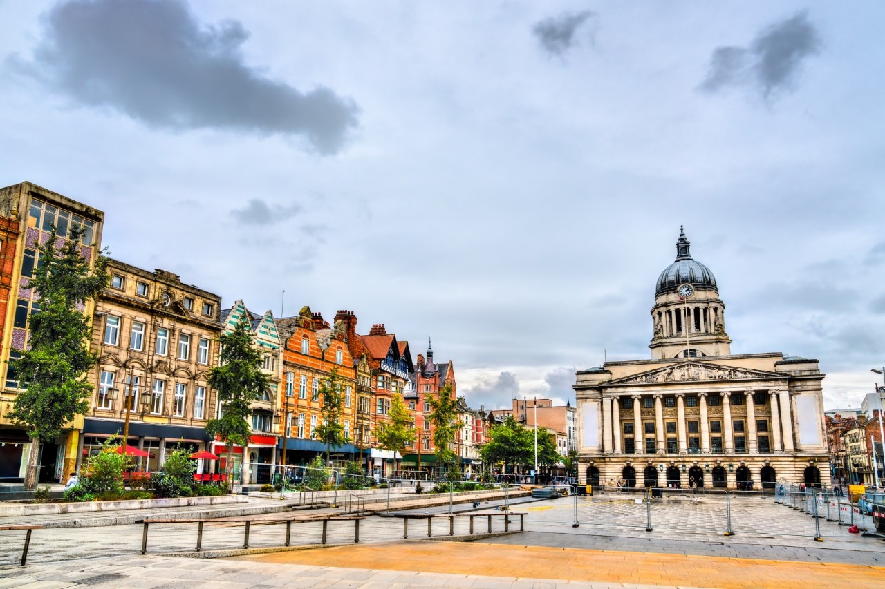 The image shows a central square with historic buildings, including a magnificent dome on a neoclassical building. On the left are colourful, multi-storey houses with scattered green trees in front of them.