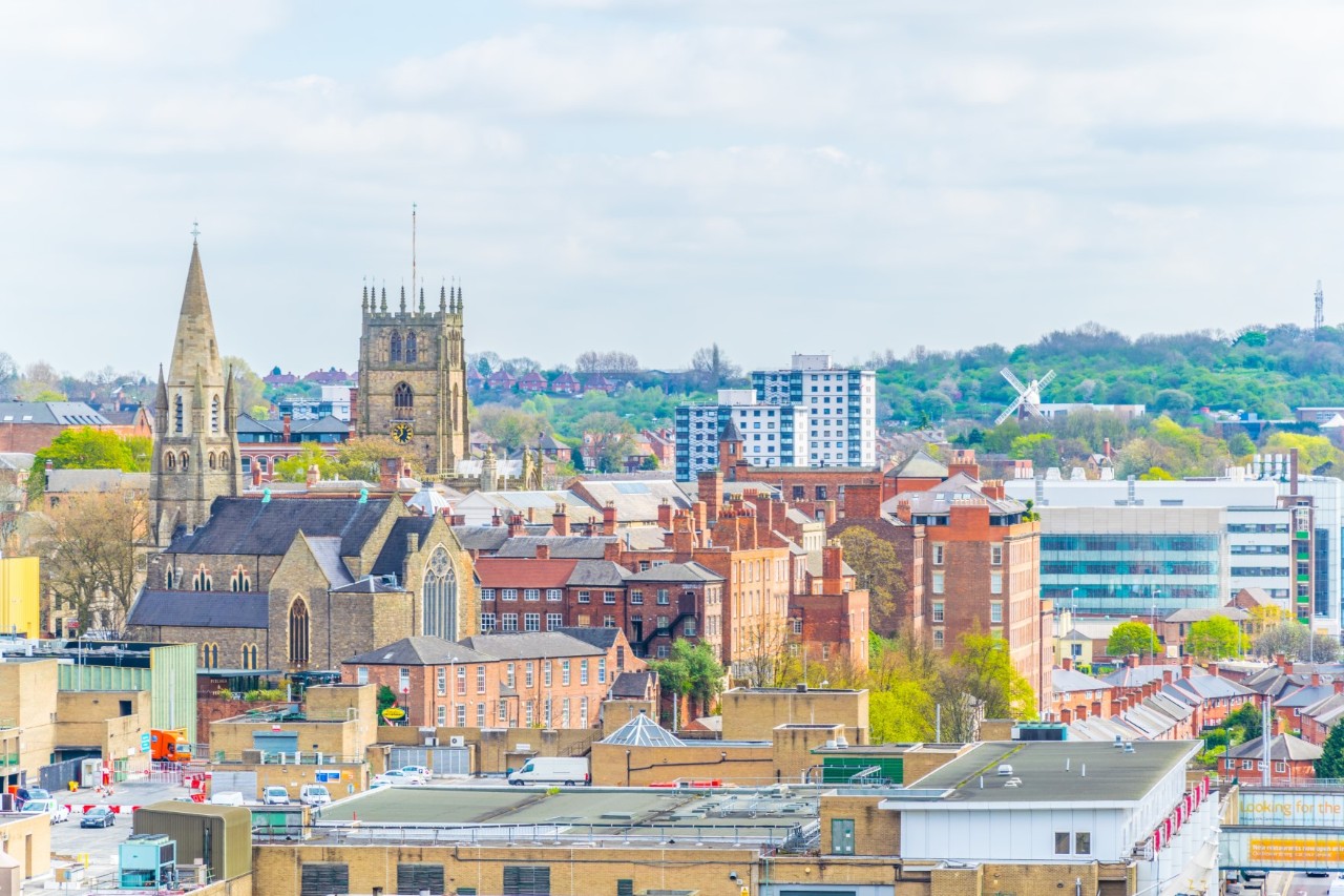The image shows an urban view with two striking churches: a neo-Gothic church with a pointed tower (left) and a larger church with a rectangular tower (right). Modern buildings and trees can be seen in the background, while the sky is overcast with clouds.