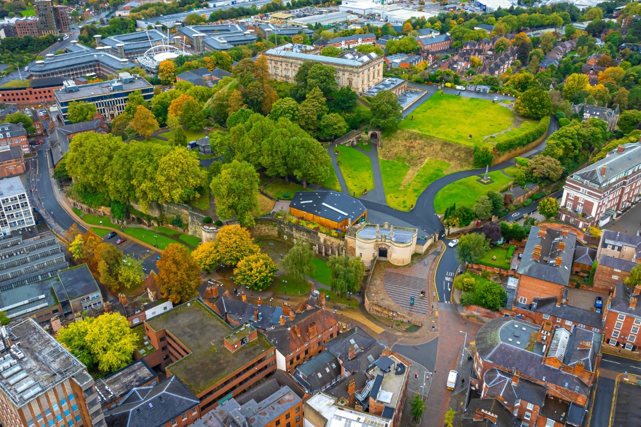 Aerial view of a castle perched on a green hill, surrounded by dense trees and parks. A town runs around the hill.