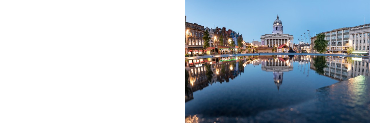 The picture shows an old market square in Nottingham at dusk. In the centre stands a magnificent building with a striking dome and a tympanum with columns below. The square is surrounded by historic and modern buildings, which are reflected in the water of a large basin in the centre.