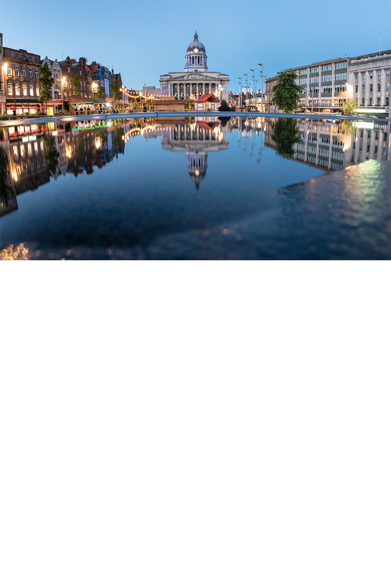 The picture shows an old market square in Nottingham at dusk. In the centre stands a magnificent building with a striking dome and a tympanum with columns below. The square is surrounded by historic and modern buildings, which are reflected in the water of a large basin in the centre.