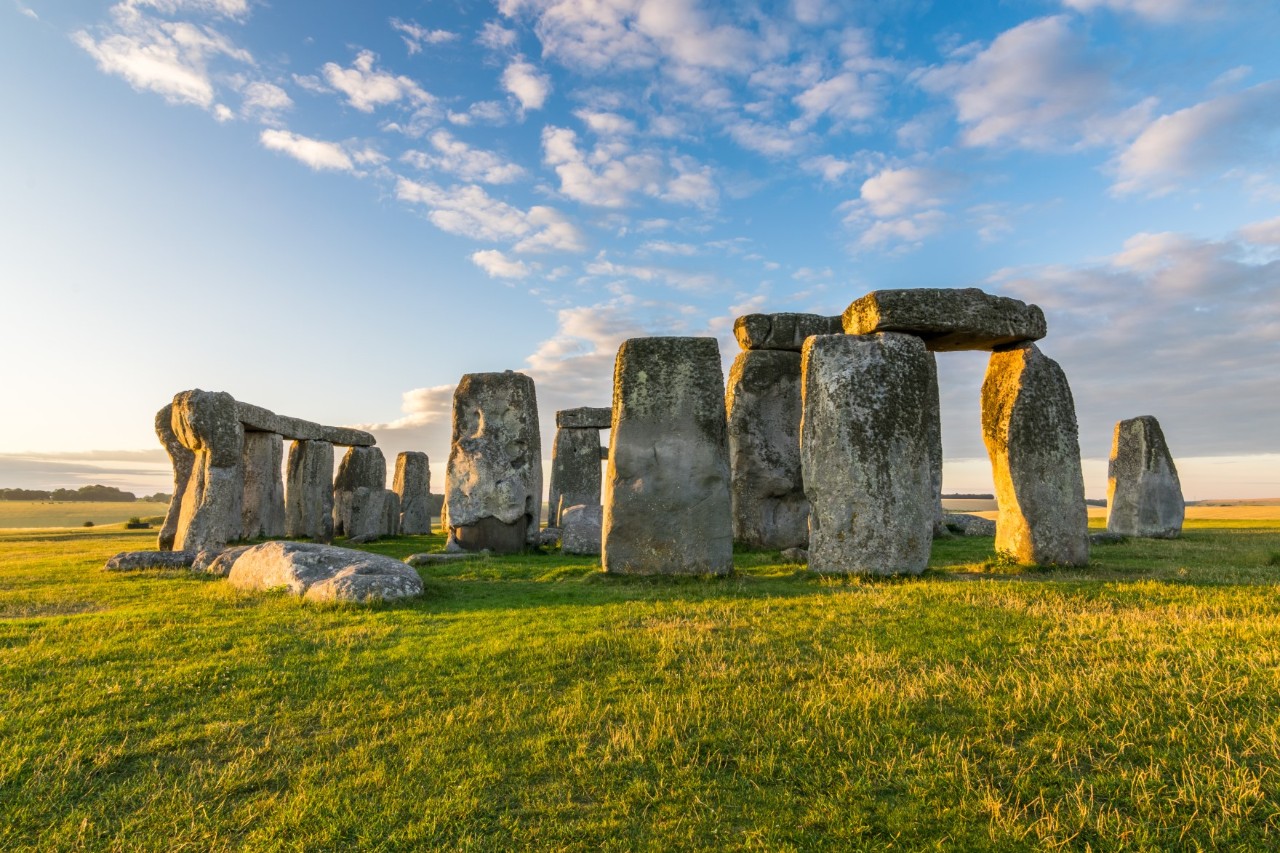 The picture shows an arrangement of massive, upright stones, some of which are bridged by horizontally lying stones. The huge boulders are surrounded by a green meadow, the landscape in the background is flat and expansive.
