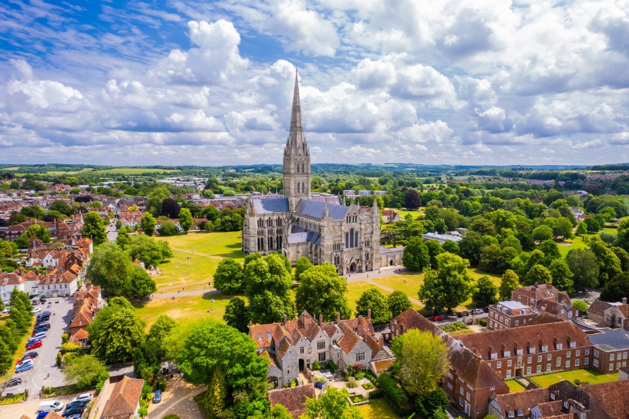 The picture shows an aerial view of an imposing cathedral with a striking tower in the middle of a green landscape, surrounded by trees, meadows and old houses.