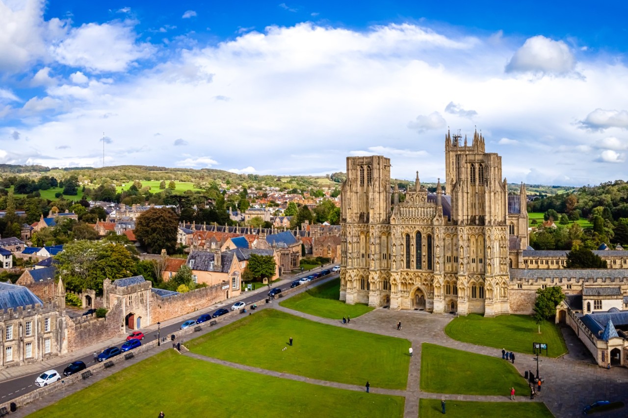 The picture shows a Gothic cathedral with elaborate decorations. It is surrounded by well-kept green spaces and old houses in a rural setting.