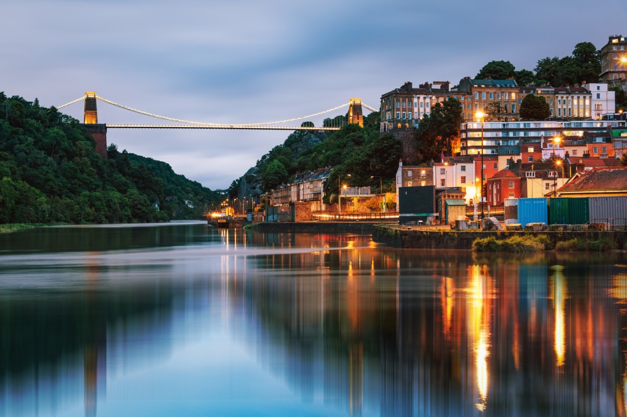 The image shows a large, illuminated bridge spanning a calm river. There are houses and lights on the right bank, while a wooded hill complements the image on the left.