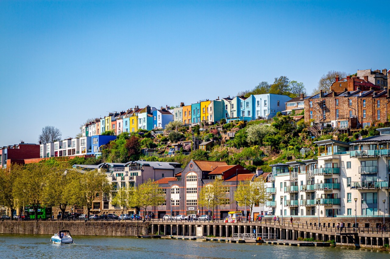 The picture shows a row of colourful houses on a hill above a river. On the bank, there are modern apartment buildings with balconies. People can be seen walking on a footbridge and a small boat is sailing on the river.