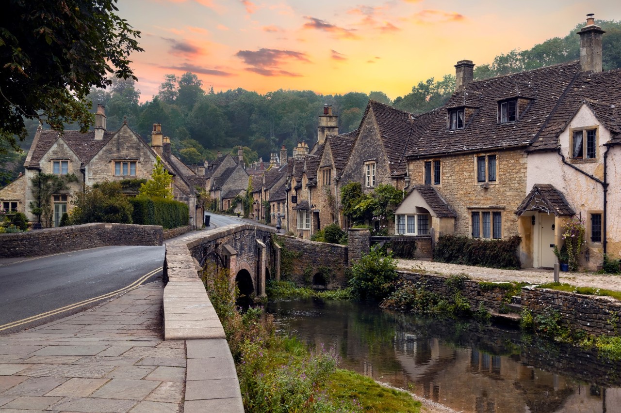 The picture shows an idyllic village with traditional English stone houses stretching along a small shallow river. A bridge crosses the water. The village is surrounded by woodland and the sky is bathed in warm colours