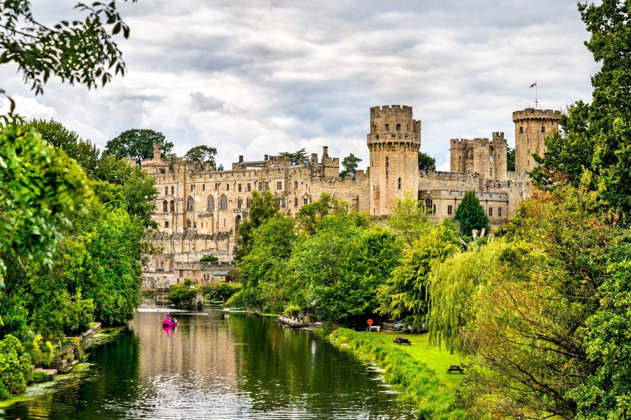 The picture shows a large medieval castle with high towers, surrounded by lush nature. A river with a small, pink-coloured boat can be seen in the foreground.