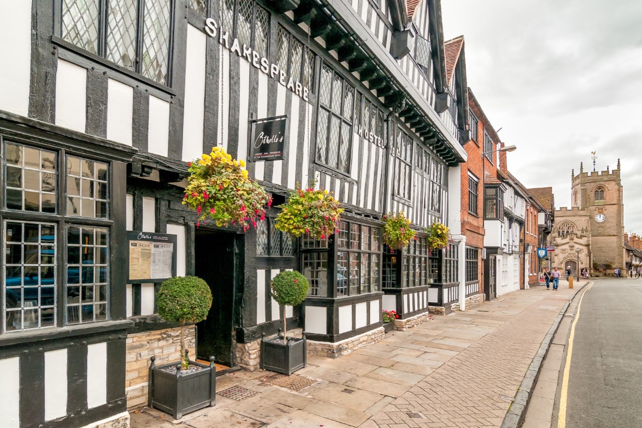 The picture shows a street with historic half-timbered houses. One of the buildings is black and white, bears the lettering "Shakespeare" and is decorated with hanging baskets of flowers. There is a church in the background