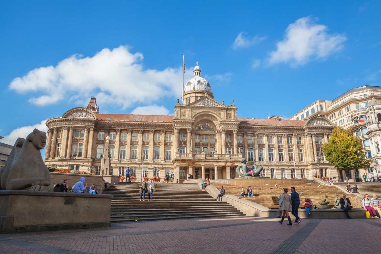 The picture shows a large Victorian house with an impressive façade and cupola. In the foreground are wide steps with people sitting on them. A large sculpture can be seen on the left.