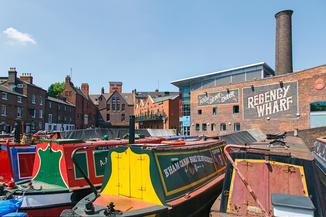 The picture shows several colourful canal boats. Old brick buildings can be seen in the background. A larger building with a modern extension and chimney bears the lettering "Regency Wharf".