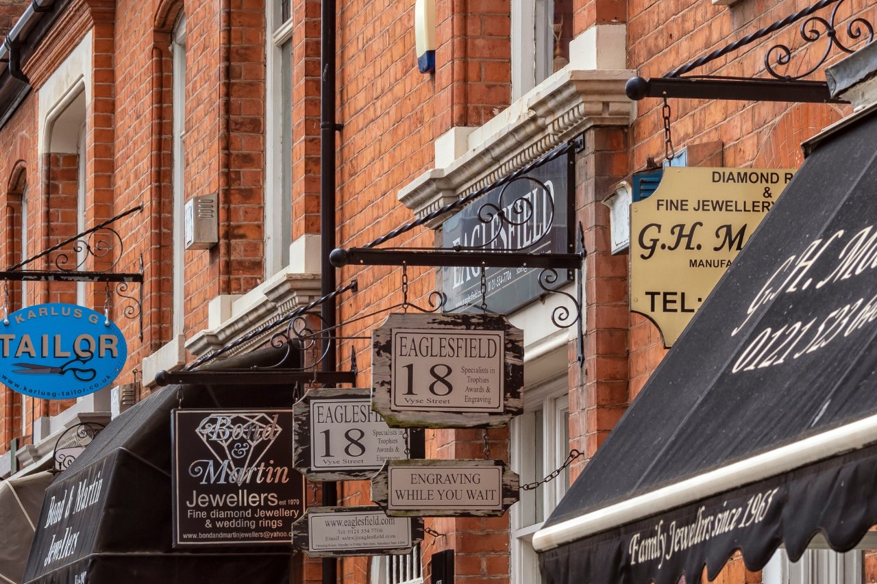 The picture shows house facades with signs from jewellers and tailors. These are attached to wrought-iron brackets on red brick houses.