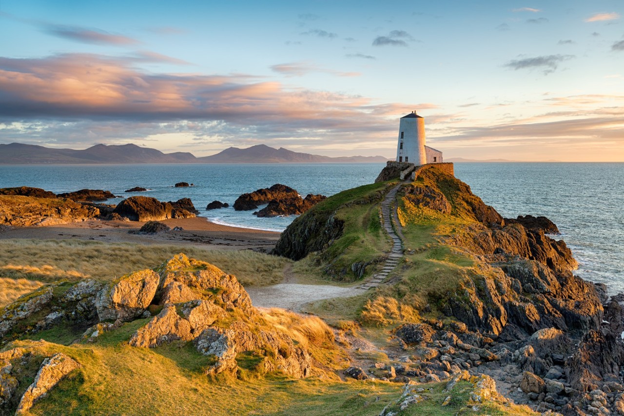 The image shows a white lighthouse on a hill, part green and part rocky, which can be reached by a narrow path. In the background, there is a sea, a coastline, and mountains.