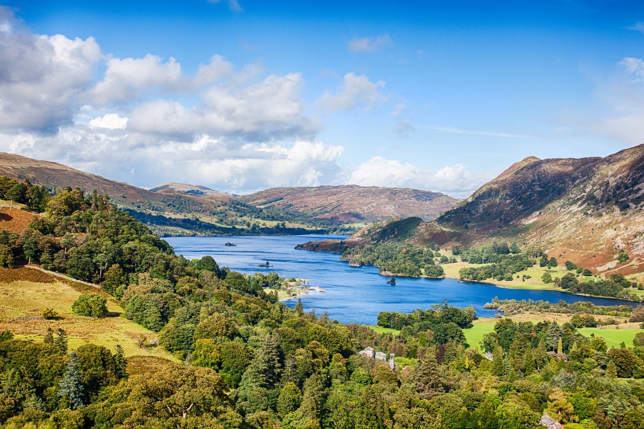 The picture shows a landscape with a calm lake, surrounded by green forests, rolling hills and mountains.