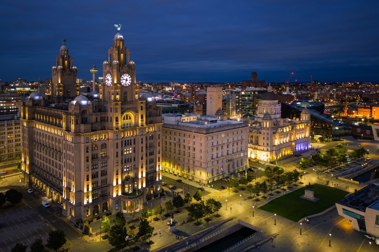 The image shows a cityscape at night, with illuminated historical buildings, including a striking building with two tower clocks, surrounded by a lively square and a canal in the foreground.