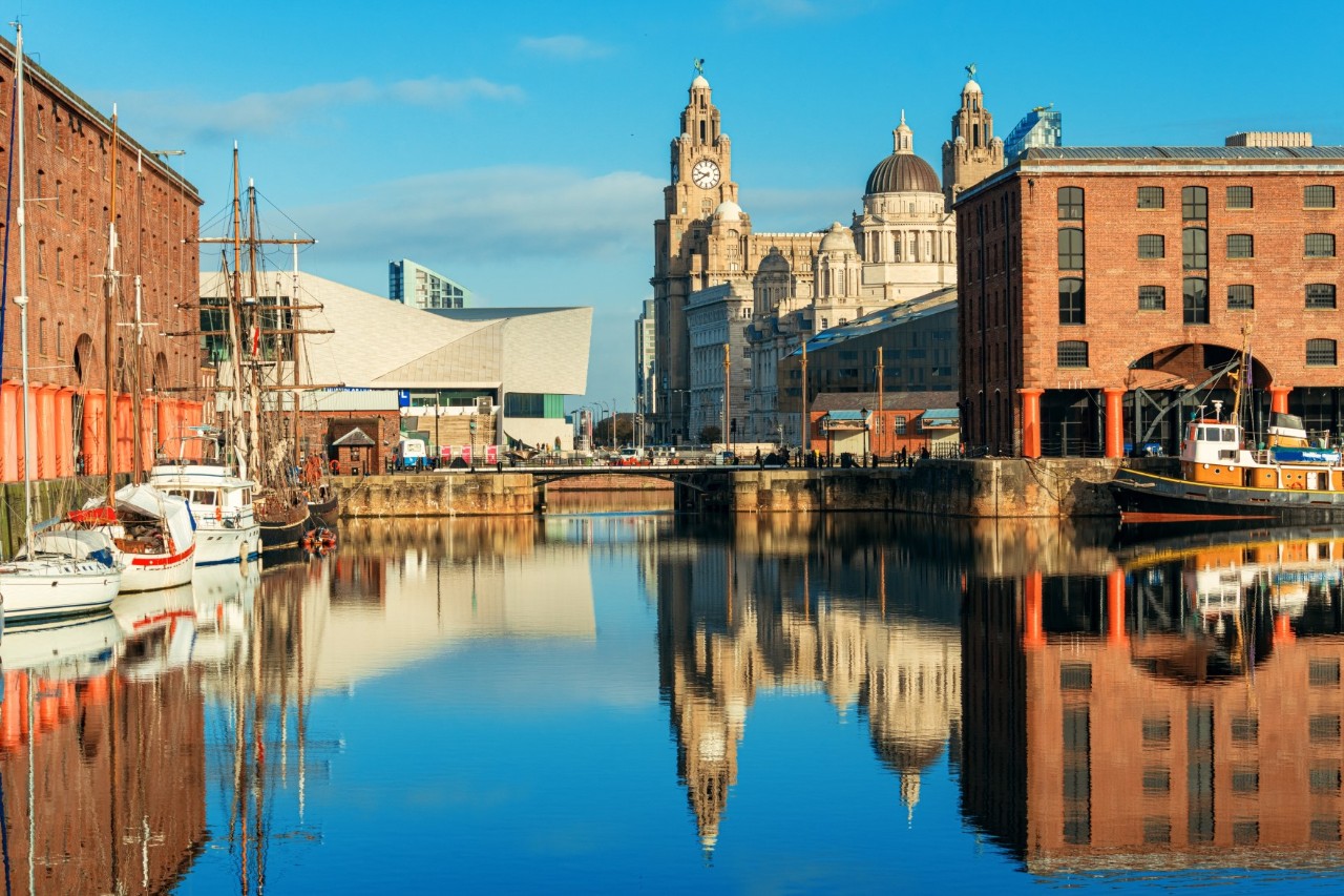 The picture shows a harbour area with historic buildings made of red brick, sailing boats in the water, and a prominent building with towers in the background. The houses and boats are reflected in the calm water of the harbour.