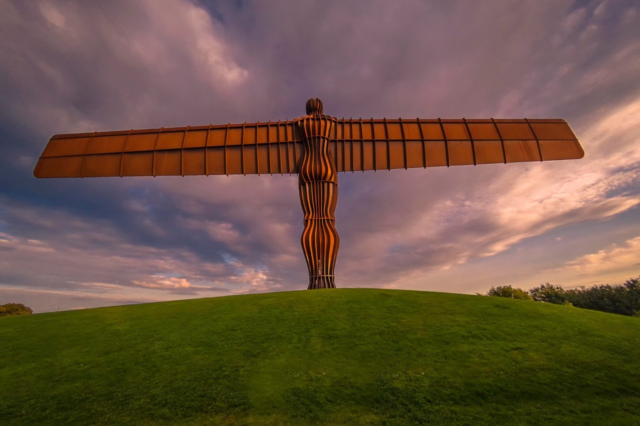 A large, human-shaped sculpture made of reddish, shimmering metal, spreading its great wings, stands on a green hill.