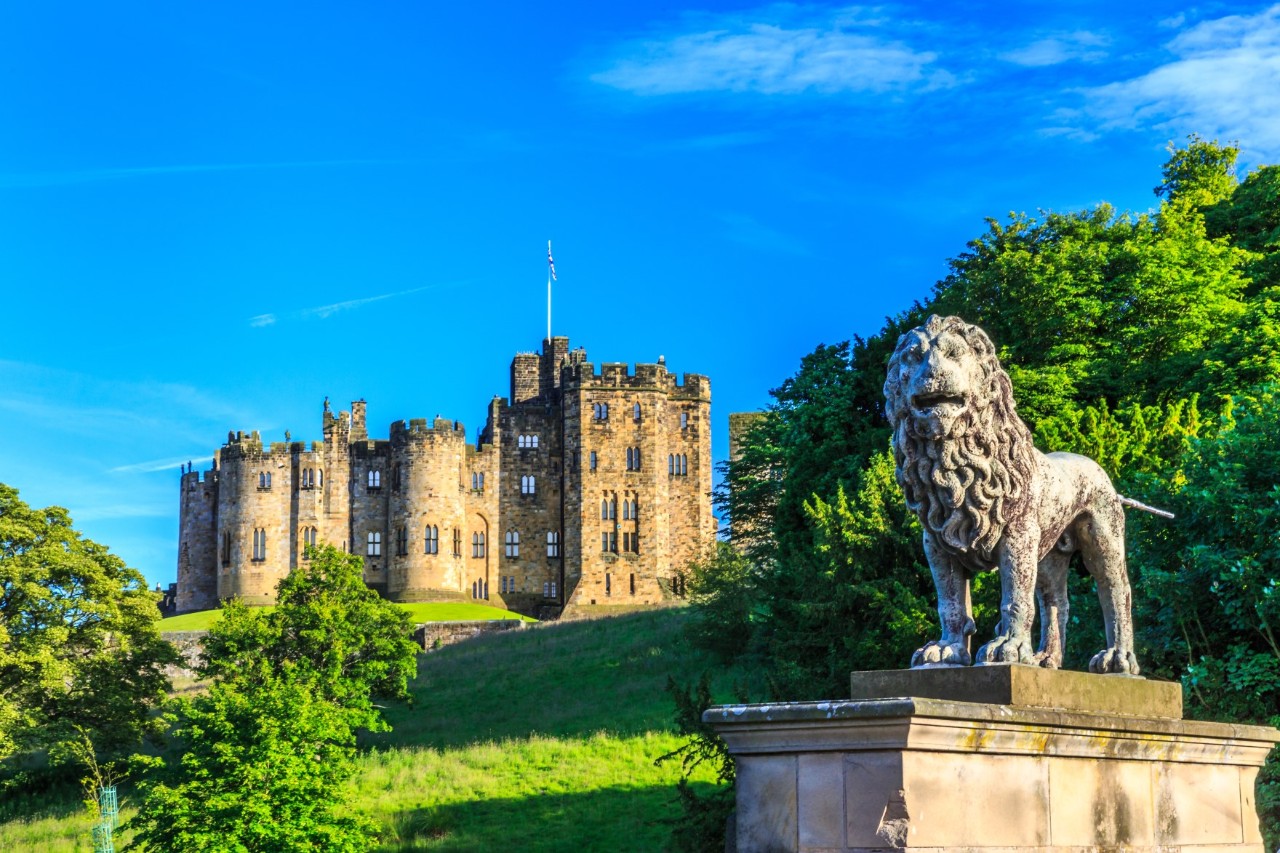 Large, old, sandstone-coloured castle, standing in a large, hilly, green garden. In the foreground, a light-coloured lion sculpture is visible on a stone pedestal.